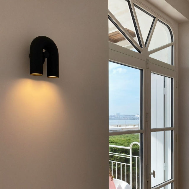 "A black, contemporary wall lamp with a looped design, mounted on a white wall next to a large window with a view."
