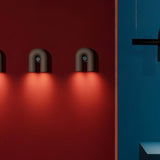 "Three black, contemporary wall lamps with a looped design, mounted on a vibrant red wall, emitting a warm glow."