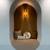 "A gray, contemporary wall lamp with a looped design, illuminating a cozy alcove with a built-in seat and decorative pillows."