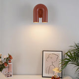 "A red, contemporary wall lamp with a looped design, mounted on a white wall above a side table with decorations."