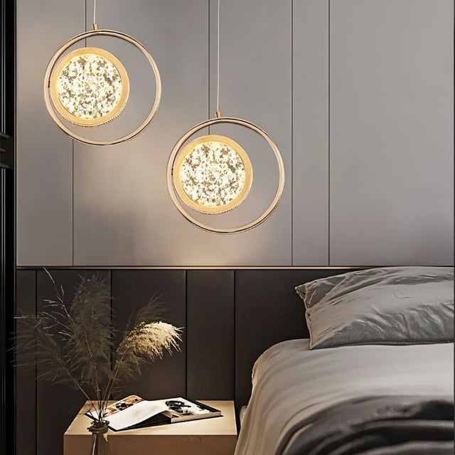 "Two gold ring pendant lights with crystal-like centers, hanging above a nightstand and bed in a room with a gray paneled wall."