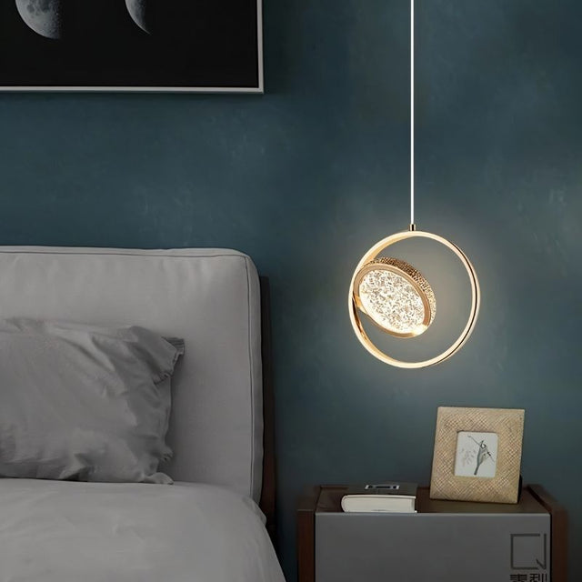 "A gold ring pendant light with a crystal-like center, hanging next to a bed in a room with a dark blue wall."