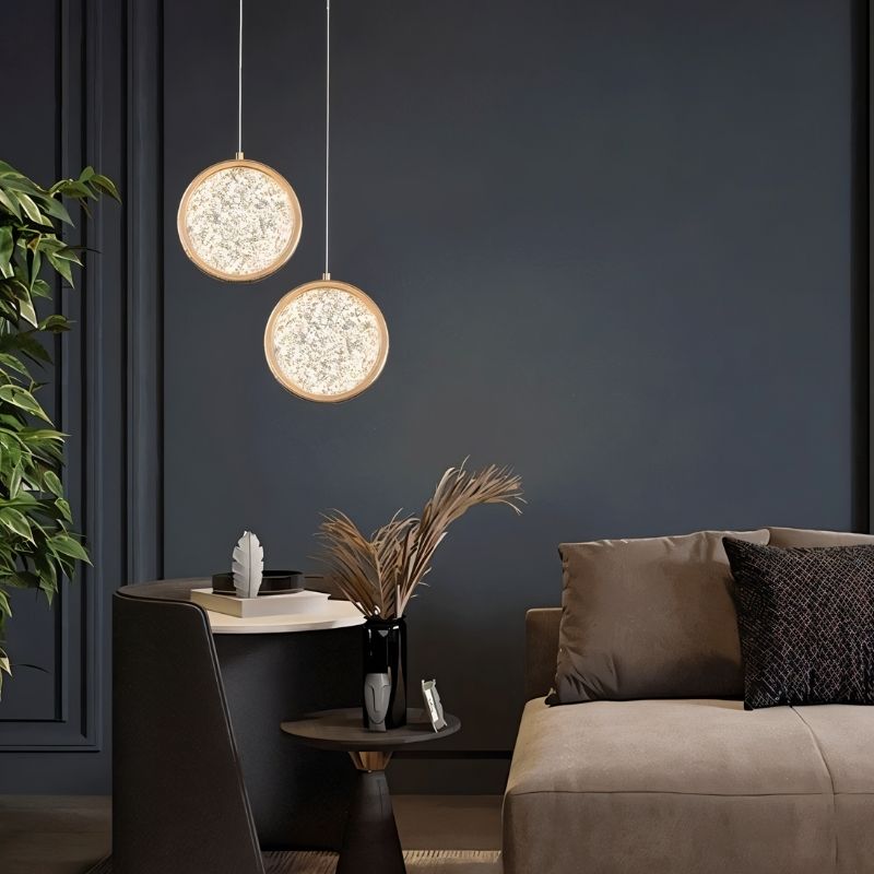 "Two gold ring pendant lights with crystal-like centers, hanging in a living room with a dark blue wall and brown furniture."