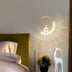 Gold crescent moon pendant light with seated astronaut figure, illuminated above bedroom nightstand.