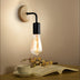 Wall-mounted lamp with natural wood backplate, black metal arm, and vintage Edison-style bulb above a reading desk with book and coffee cup.