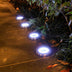 Four round stainless steel in-ground lights with bright white LED glow installed along a garden pathway at night.