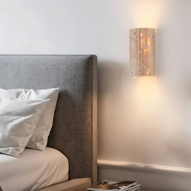 "A minimalist, rectangular travertine wall lamp mounted on a bedroom wall, providing a soft, ambient glow next to a bed at night."