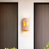 "A minimalist, rectangular travertine wall lamp mounted on a white exterior wall between two dark doorways, providing a soft, ambient glow."