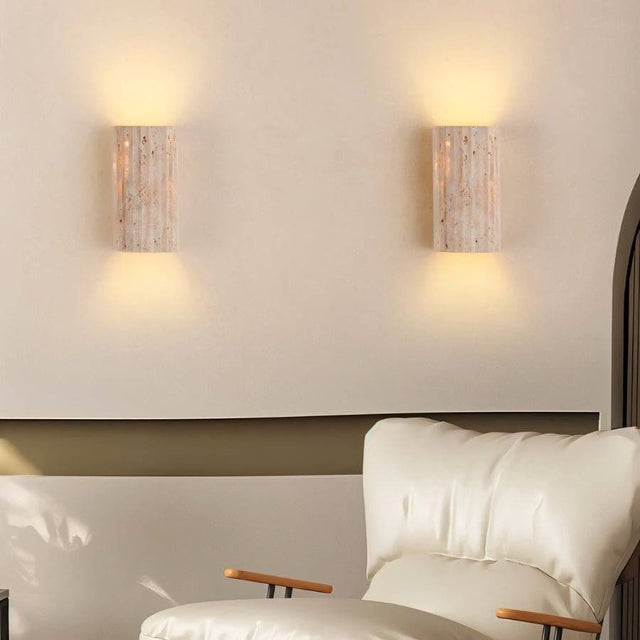 "Pair of minimalist, rectangular travertine wall lamps mounted symmetrically on a living room wall, providing up and down lighting, with a cream-colored armchair in the foreground."