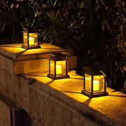 Three black solar lanterns with glowing yellow lights arranged on a concrete ledge against dark foliage.