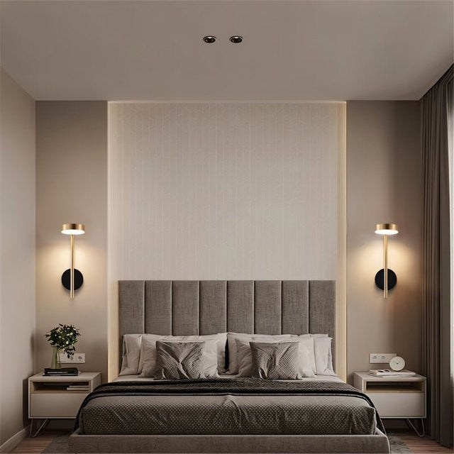 "Two gold and black wall lamps with round bases and vertical light sources, installed on either side of a gray tufted headboard in a bedroom with a textured wall and gray curtains."