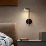 "A gold and black wall lamp with a round base and a vertical light source, installed above a nightstand with books, next to a gray tufted headboard in a bedroom."