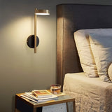 "A gold and black wall lamp with a round base and a vertical light source, installed above a nightstand with books and a glass of water, next to a gray tufted headboard in a bedroom."