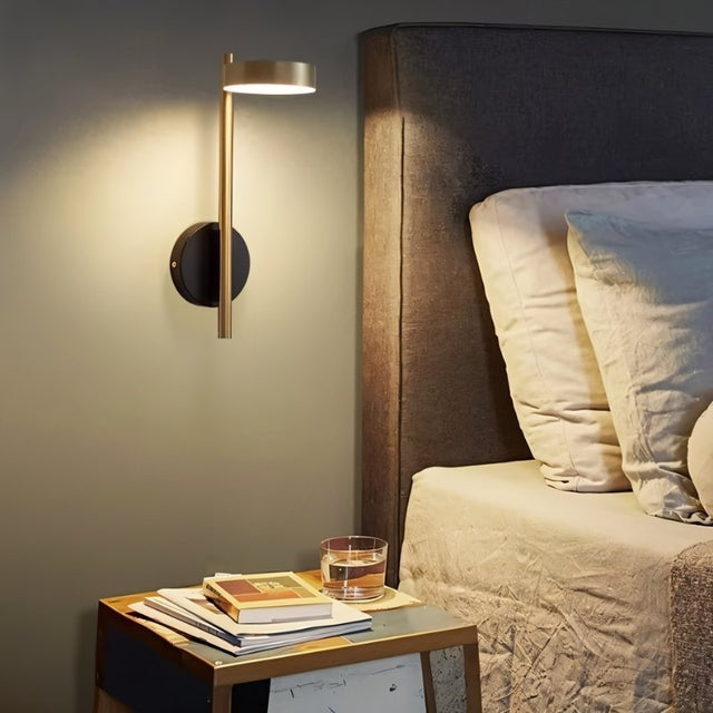 "A gold and black wall lamp with a round base and a vertical light source, installed above a nightstand with books and a glass of water, next to a gray tufted headboard in a bedroom."