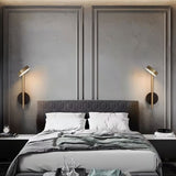 "Two gold and black wall lamps with round bases and vertical light sources, installed on either side of a gray tufted headboard in a bedroom."