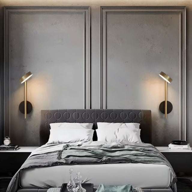 "Two gold and black wall lamps with round bases and vertical light sources, installed on either side of a gray tufted headboard in a bedroom."
