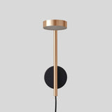 "A gold and black wall lamp with a round base and a vertical light source, shown against a gray background."