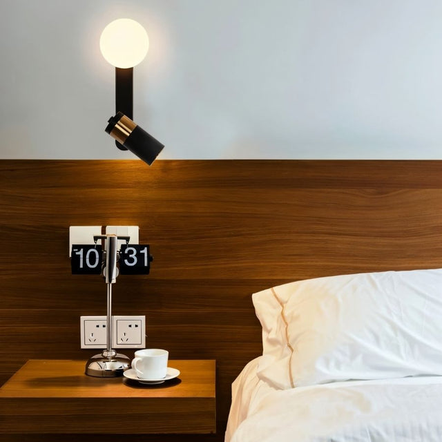 "A modern black bedside reading lamp with frosted glass globe and adjustable spotlight, mounted on a white wall above a wooden headboard, alongside a flip clock on a bedside table."