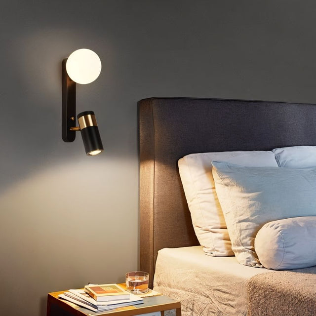 "A modern black bedside reading lamp with a frosted glass globe and adjustable spotlight, mounted on a gray wall above a bed with a gray headboard."