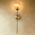 Brass wall sconce with clear glass sphere shade and long stem against gray wall, lifestyle setting with bed and plant.