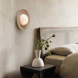 "Modern, minimalist wall mount light with round travertine base and frosted globe on bedroom wall above side table with vase and floral arrangement."