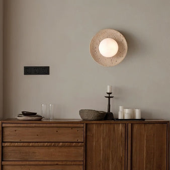 "Modern, minimalist wall mount light with a round travertine base and frosted globe, mounted on a living room wall above a wooden console table with decorative items."