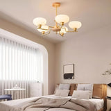 "Brass and wood chandelier with five white globe-shaped glass shades, hanging in a modern bedroom with a large window and artwork."