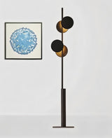 "A modern floor lamp with sculptural, offset circular shades, standing next to a framed artwork on a white wall."