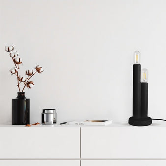 "A minimalist, two-light black table lamp on a white cabinet in a modern, minimalist home setting with a decorative vase and a book."