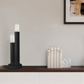 "A minimalist, two-light black table lamp providing soft illumination in a modern living room with books and artwork."