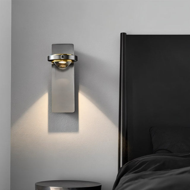 "A modern gunmetal wall lamp with a glass lens illuminating a bedroom wall next to a dark headboard."