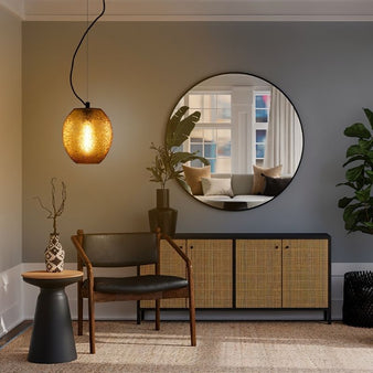 "An amber glass pendant lamp with a textured, organic design hanging in a modern living room with a round mirror, a wooden chair, and a woven cabinet."