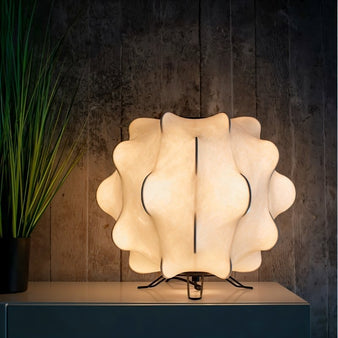 "A white table lamp with a cloud-like shade emitting a soft glow on a nightstand against a dark textured wall."