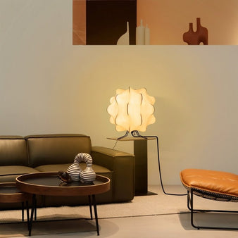 "A white table lamp with a cloud-like shade sits on a small table in a modern living room with a green sofa, artwork, and a leather ottoman."