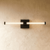 Gold brass linear vanity light with frosted glass tubes above circular gold mirror in white bathroom.