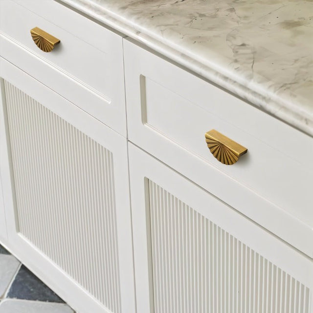 "Vintage brass fan-shaped drawer pulls with a ridged, pleated surface, installed on white bathroom drawers with grooved detailing, adding a touch of Art Deco elegance to the space."