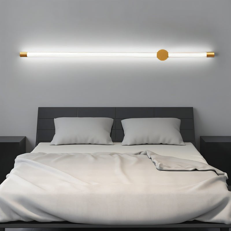 "A long, horizontal wall lamp with a brass finish and acrylic diffuser mounted above a bed in a modern bedroom, providing soft, ambient light."