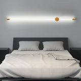 "A long, horizontal wall lamp with a brass finish and acrylic diffuser mounted above a bed in a modern bedroom, providing soft, ambient light."