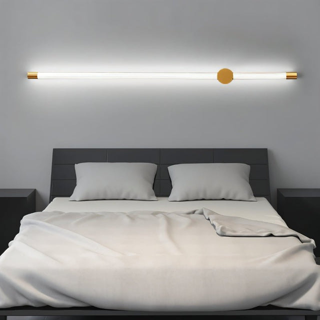 "A long, horizontal wall lamp with a brass finish and acrylic diffuser mounted above a bed in a modern bedroom, providing soft, ambient light."