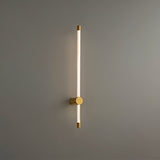 "A sleek, vertical brass wall lamp with a long, thin light bar and acrylic diffuser, mounted on a gray wall in a modern bathroom."