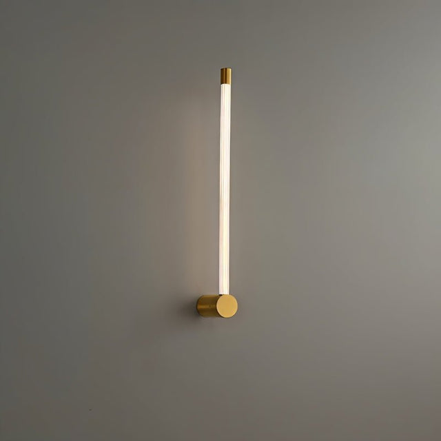"A modern brass wall lamp with a long, thin light bar and acrylic diffuser, mounted vertically on a gray wall in a bathroom setting."