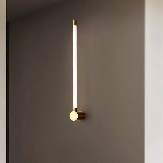 "A sleek, vertical brass wall sconce with a long, thin light bar and acrylic diffuser, mounted on a gray wall."