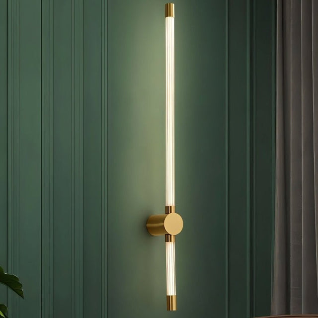 "A sleek, vertical brass wall sconce with a long, thin light bar and acrylic diffuser, mounted on a dark green textured wall."