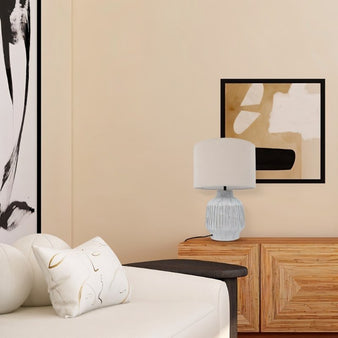 "A modern ceramic table lamp with a light-colored shade sits on a console table in a living room with abstract art on the wall."