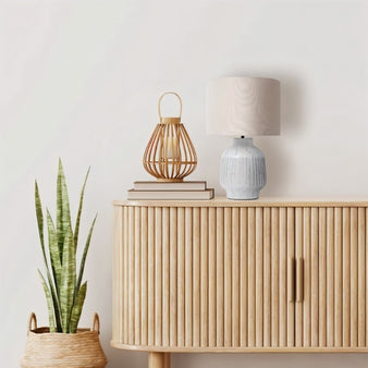 "A modern ceramic table lamp with a light-colored shade sits on a wooden sideboard next to a decorative lantern and a potted snake plant."