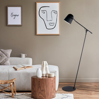 "A black floor lamp with an adjustable arm and a cone-shaped shade in a modern living room with a gray sofa, a wooden coffee table, and abstract art on the wall."