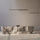"Two modern linear pendant lights hanging above a rough-textured stone table in a minimalist interior space."