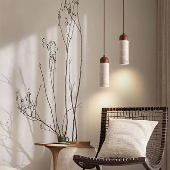 "Two cylindrical travertine Gilto pendant lights hanging above a chair and side table with dried branches."