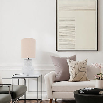 "A modern ceramic table lamp with a textured base and a light-colored shade sits on a side table in a living room with contemporary art and furniture."