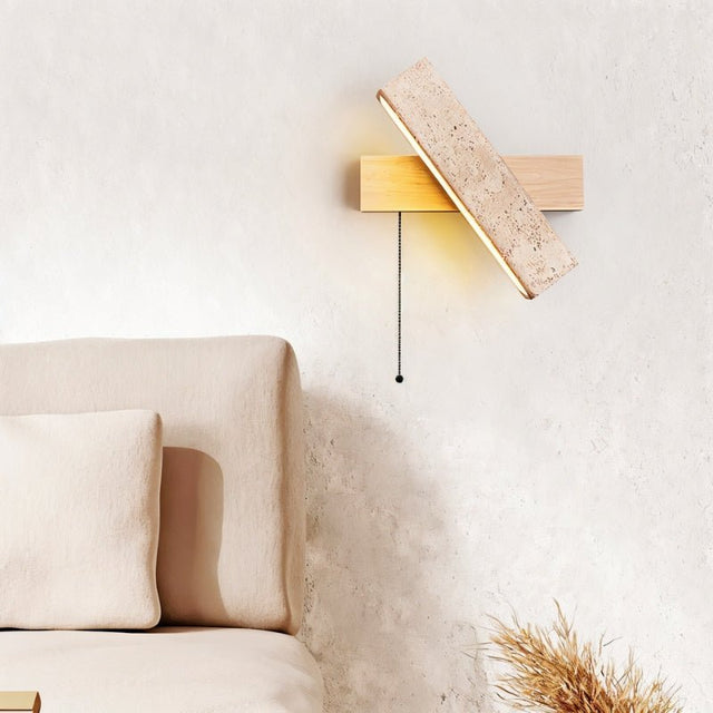 "A modern wall lamp with an angled yellow travertine shade and a light wood rectangular base, mounted on a textured wall in a bedroom, casting a warm, ambient light."